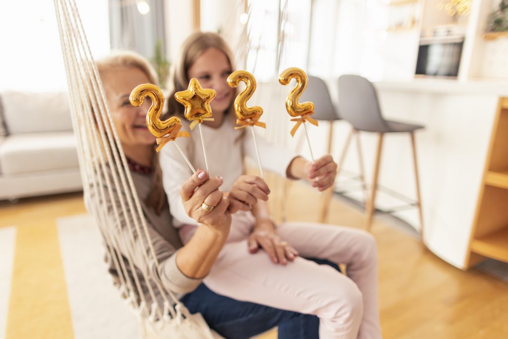 Beautiful grandmother and granddaughter sitting in hanging swing holding balloons shaped as numbers 2022 representing the upcoming New Year